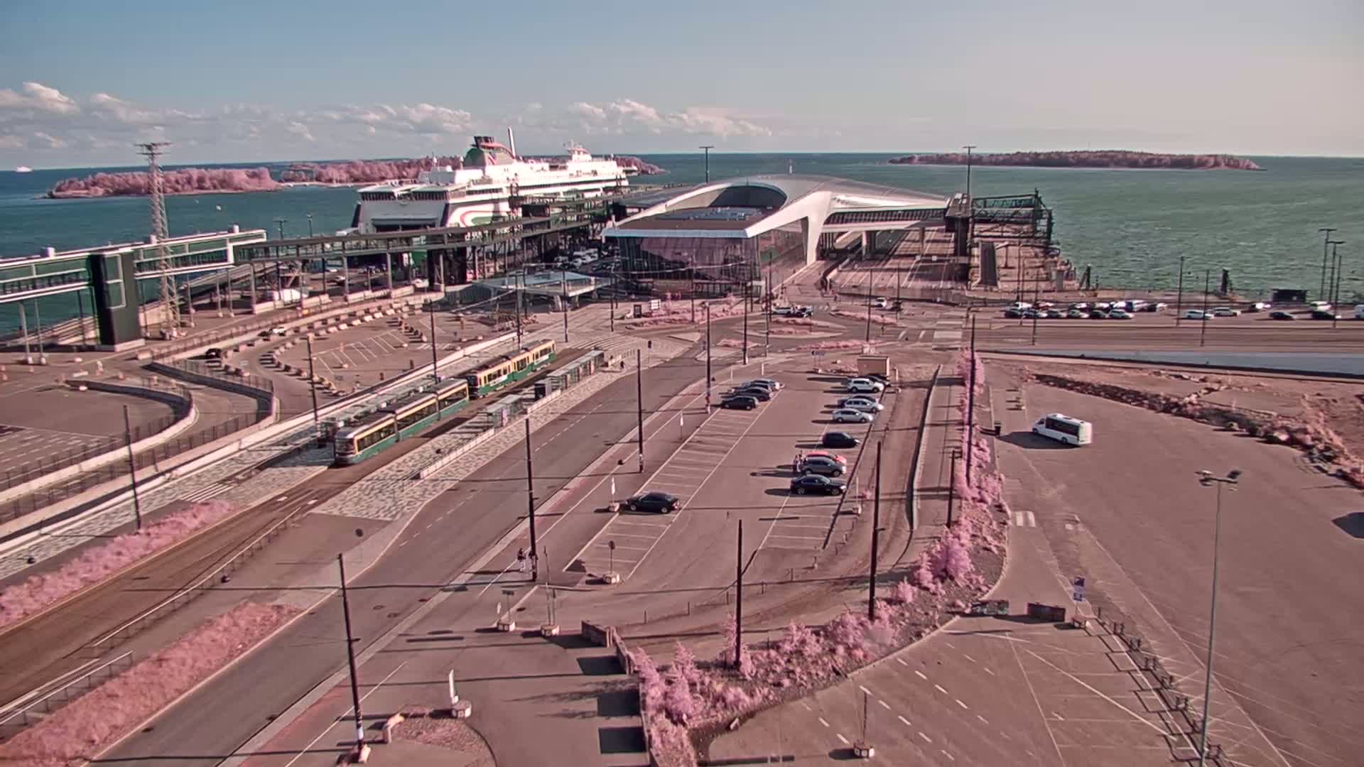 An aerial view shows a ferry terminal on a sunny day, with a ferry docked, trams at a station, cars in a parking lot, and islands visible in the distance.