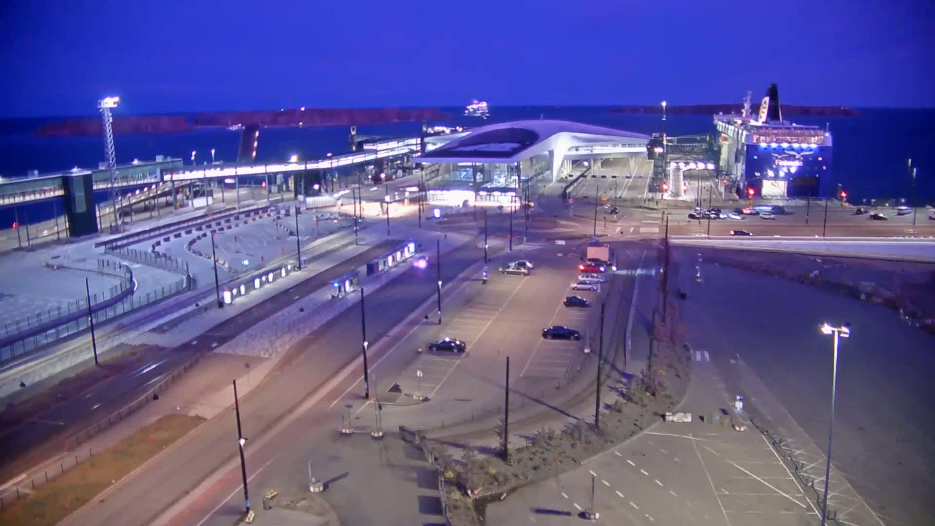 A ferry is docked at a terminal under a twilight sky, with several cars parked in a lot nearby.