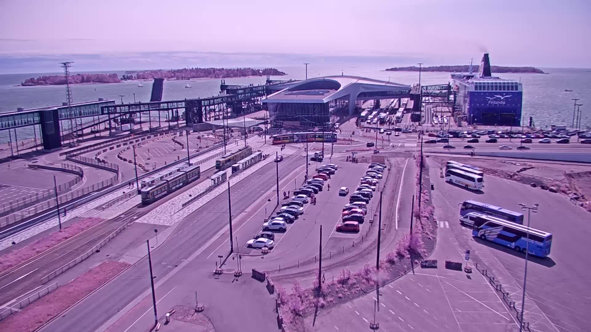 A ferry terminal on a sunny day with trams, cars, and buses parked nearby, and a large ferry docked at the terminal.