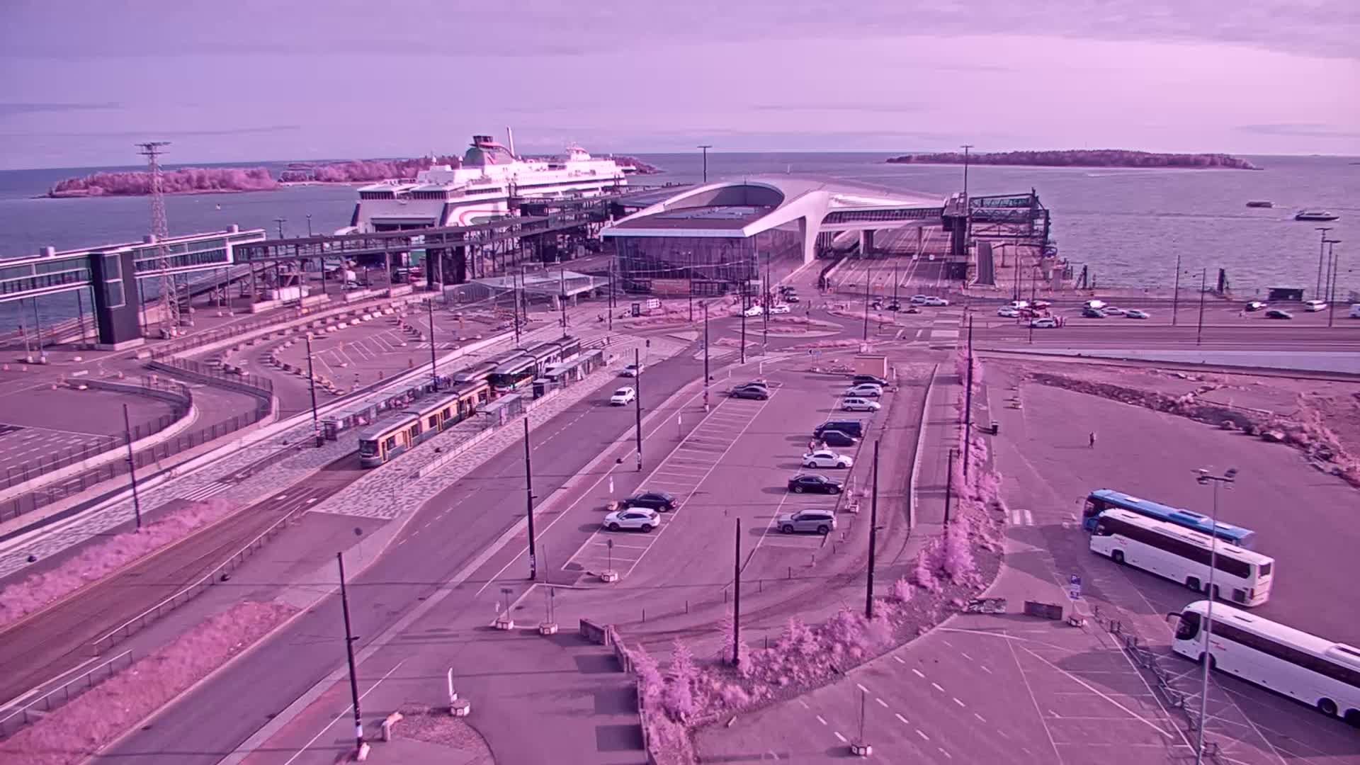 A ferry is docked at a terminal with a tram and several cars and buses in a parking lot, all under a sunny sky.