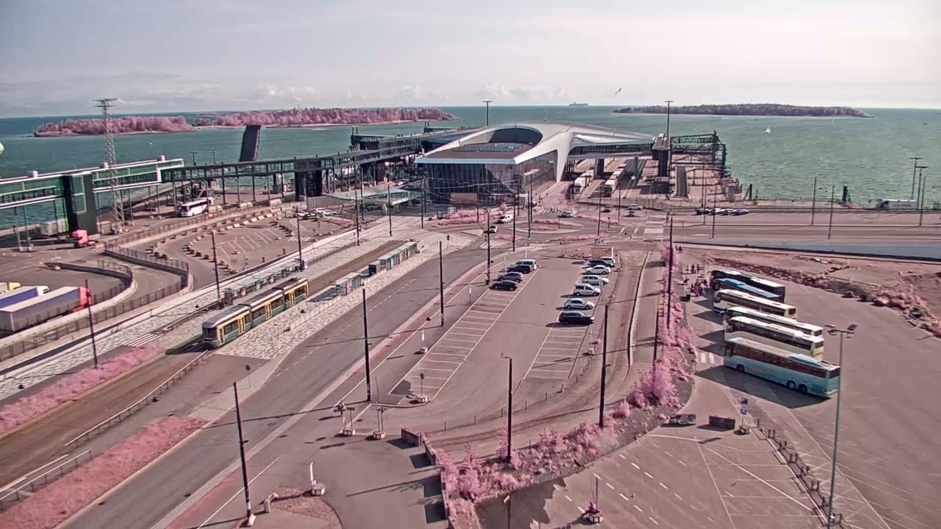 A ferry terminal with buses and a tram, a parking lot, and a body of water with islands in the background on a sunny day.