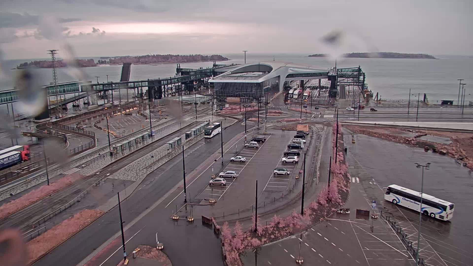 A ferry terminal with a large parking lot and several vehicles, viewed on a rainy day with overcast skies and calm water visible in the background.