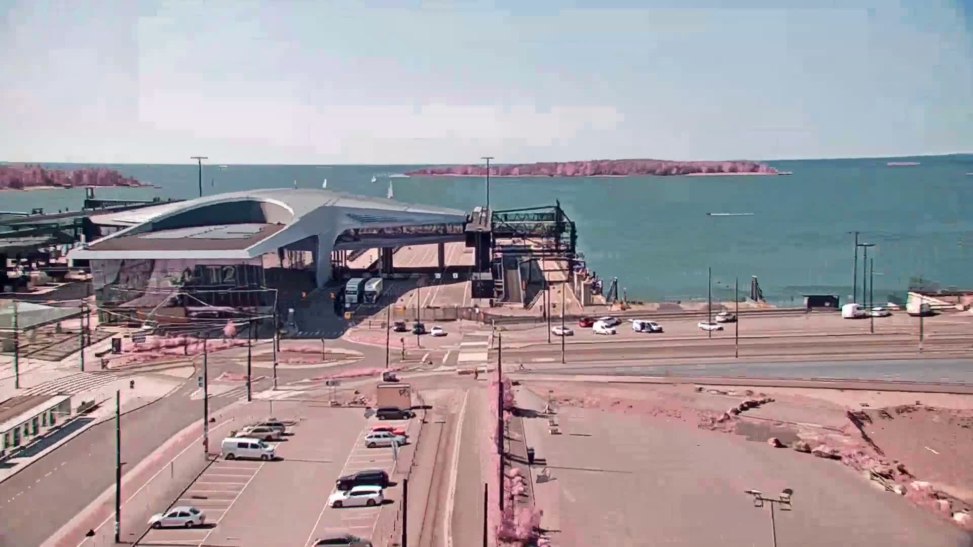 A modern ferry terminal with a curved roof sits by a calm, blue body of water on a sunny day, with several cars parked nearby and some boats visible in the distance.