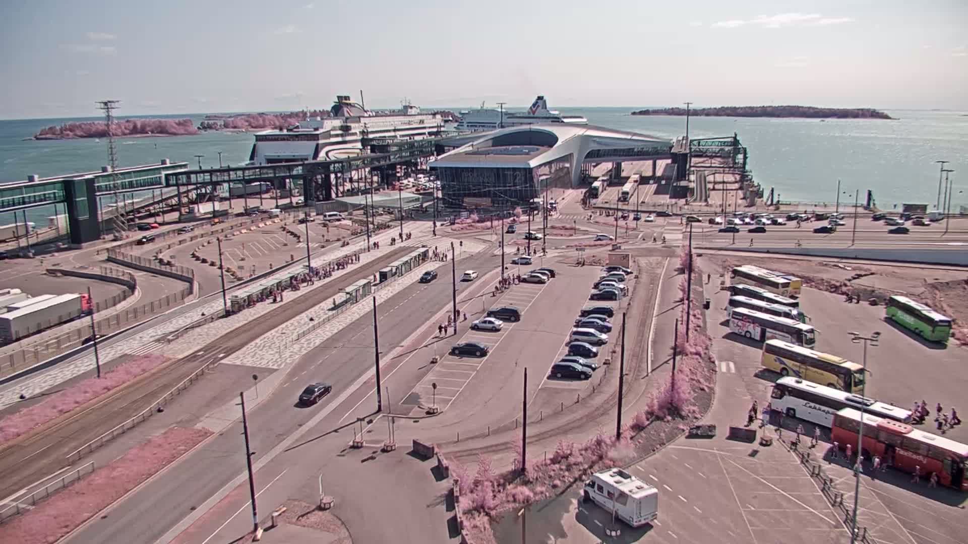 A ferry terminal on a sunny day, with several ferries docked, cars parked in lots, buses waiting, and people walking around the terminal area.