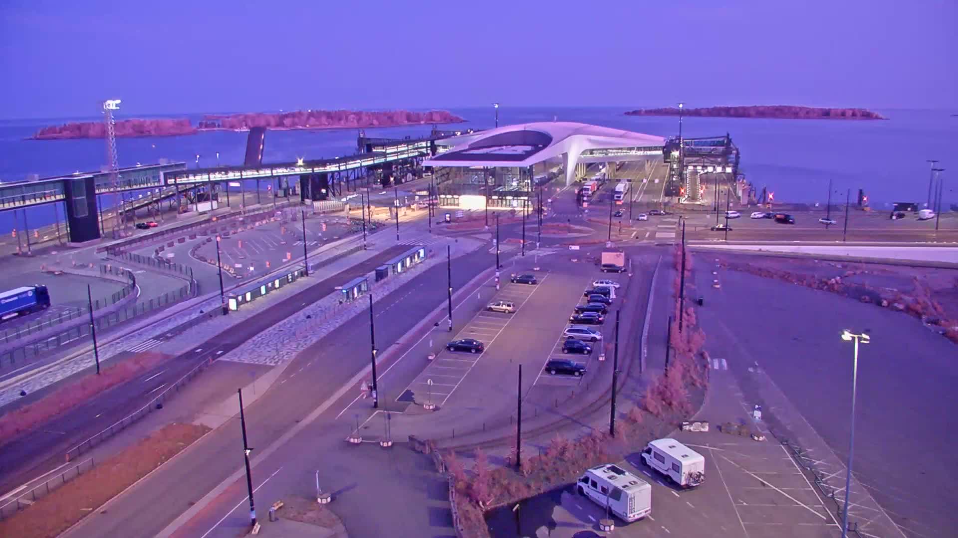 An aerial view of a ferry terminal at dusk, with cars parked nearby and a calm body of water visible beyond.