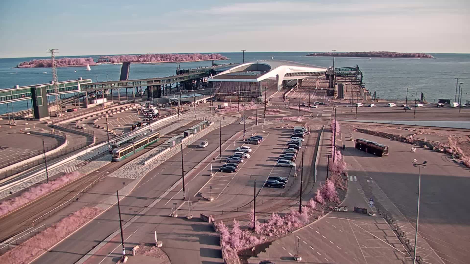 An aerial view shows a ferry terminal with a tram, cars in a parking lot, and a body of water with islands in the background under sunny skies.