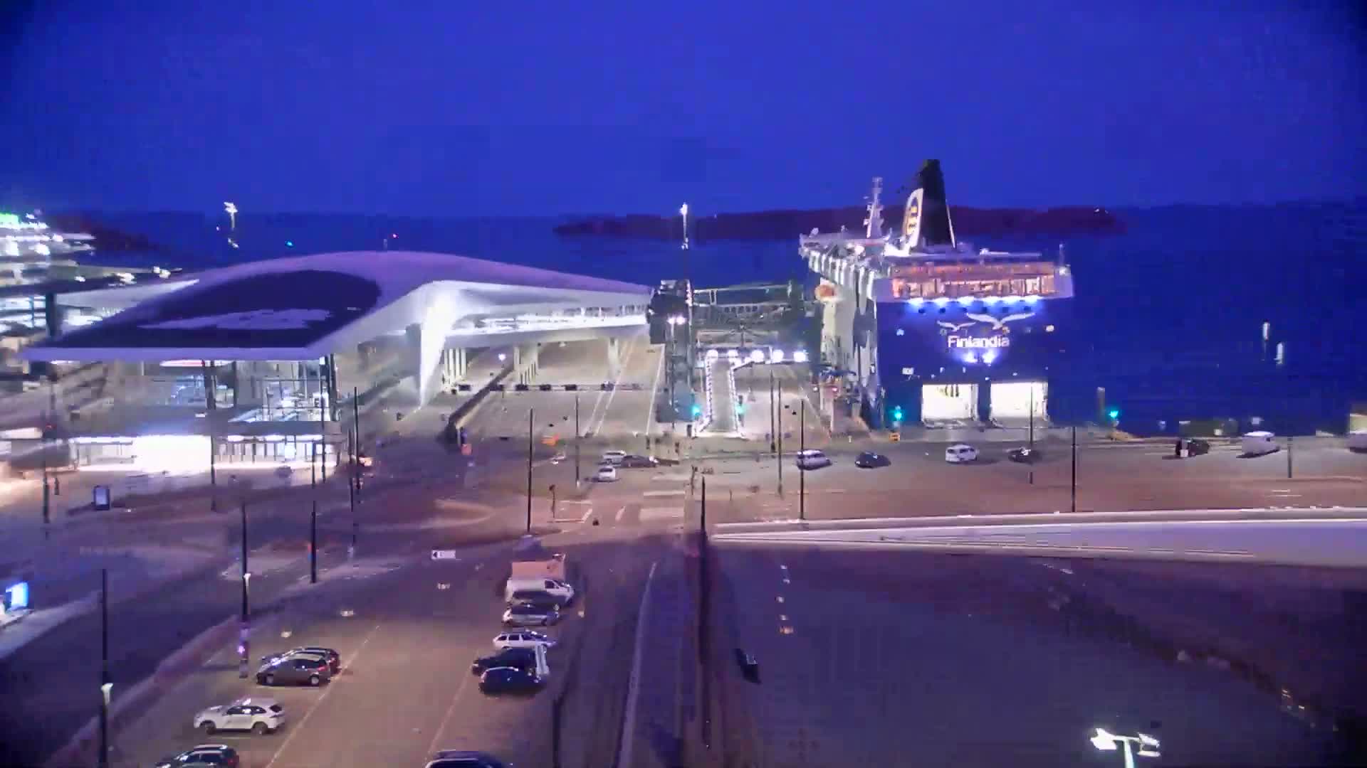 An aerial view shows a ferry terminal with a tram, cars in a parking lot, and a body of water with islands in the background under sunny skies.