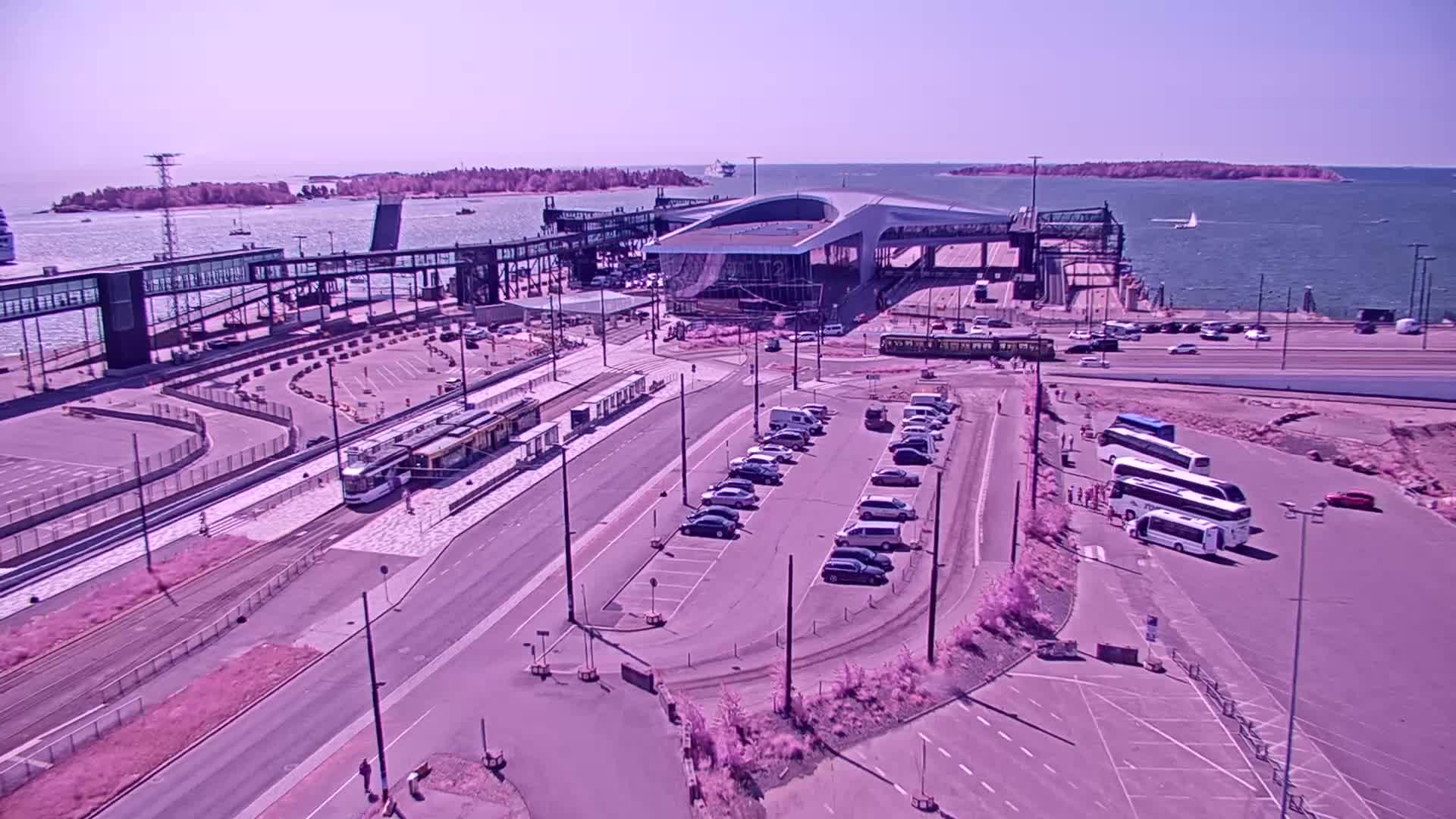 A high-angle view of a ferry terminal on a sunny day, showing a tram, cars in a parking lot, buses, and a body of water with islands in the distance.
