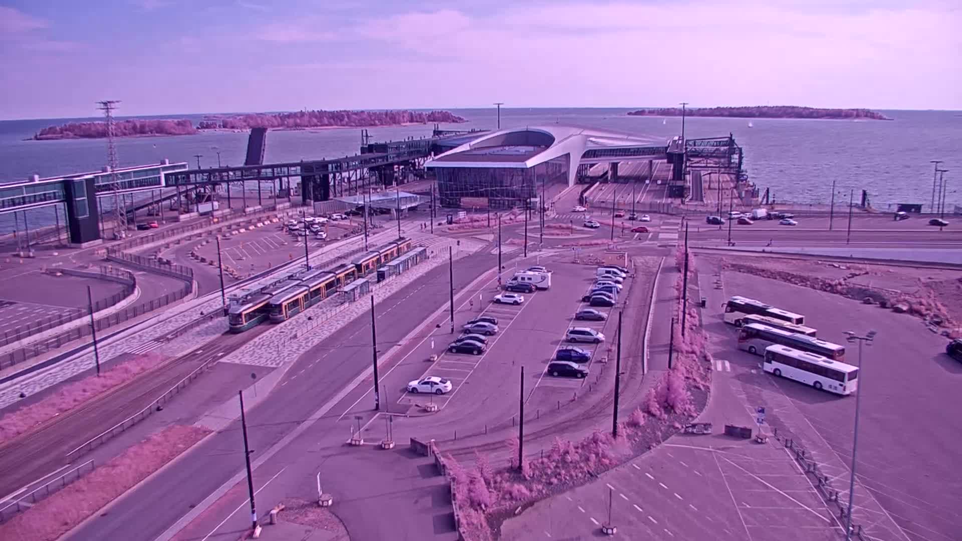 An aerial view of a ferry terminal on a sunny day shows a tram, cars in a parking lot, and several buses, with calm water and islands visible in the background.