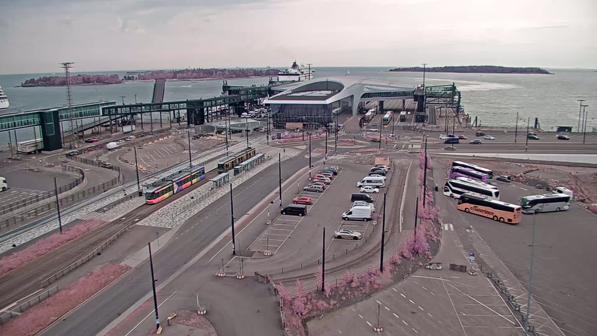 An aerial view of a ferry terminal on an overcast day shows a tram, cars in a parking lot, and several buses, with a ferry in the background and water beyond.