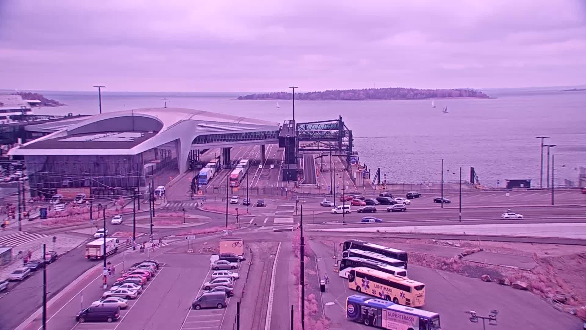 A ferry terminal with a modern building, several vehicles, and buses in a parking lot overlooks a calm body of water under a cloudy sky.