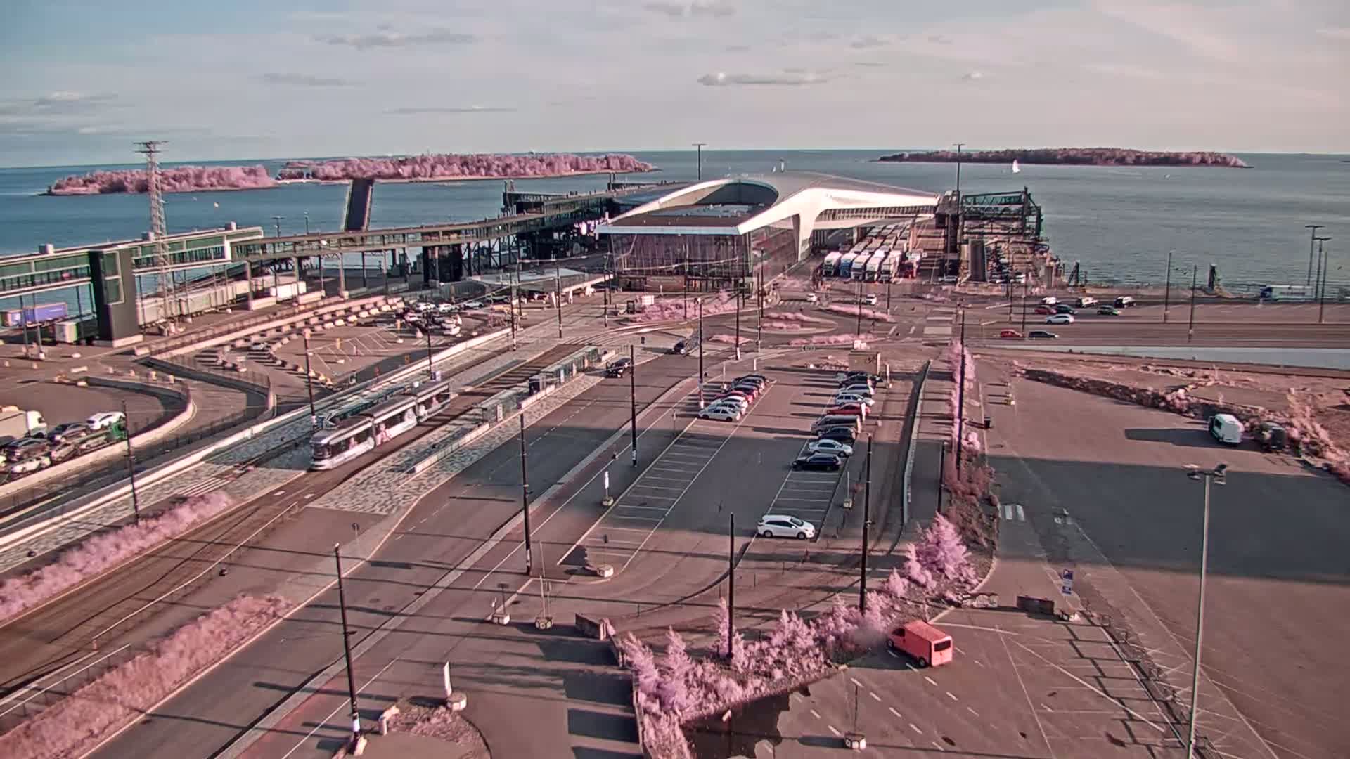 An aerial view of a ferry terminal on a partly sunny day, with a tram, cars, and trucks visible in the parking lot and surrounding area.