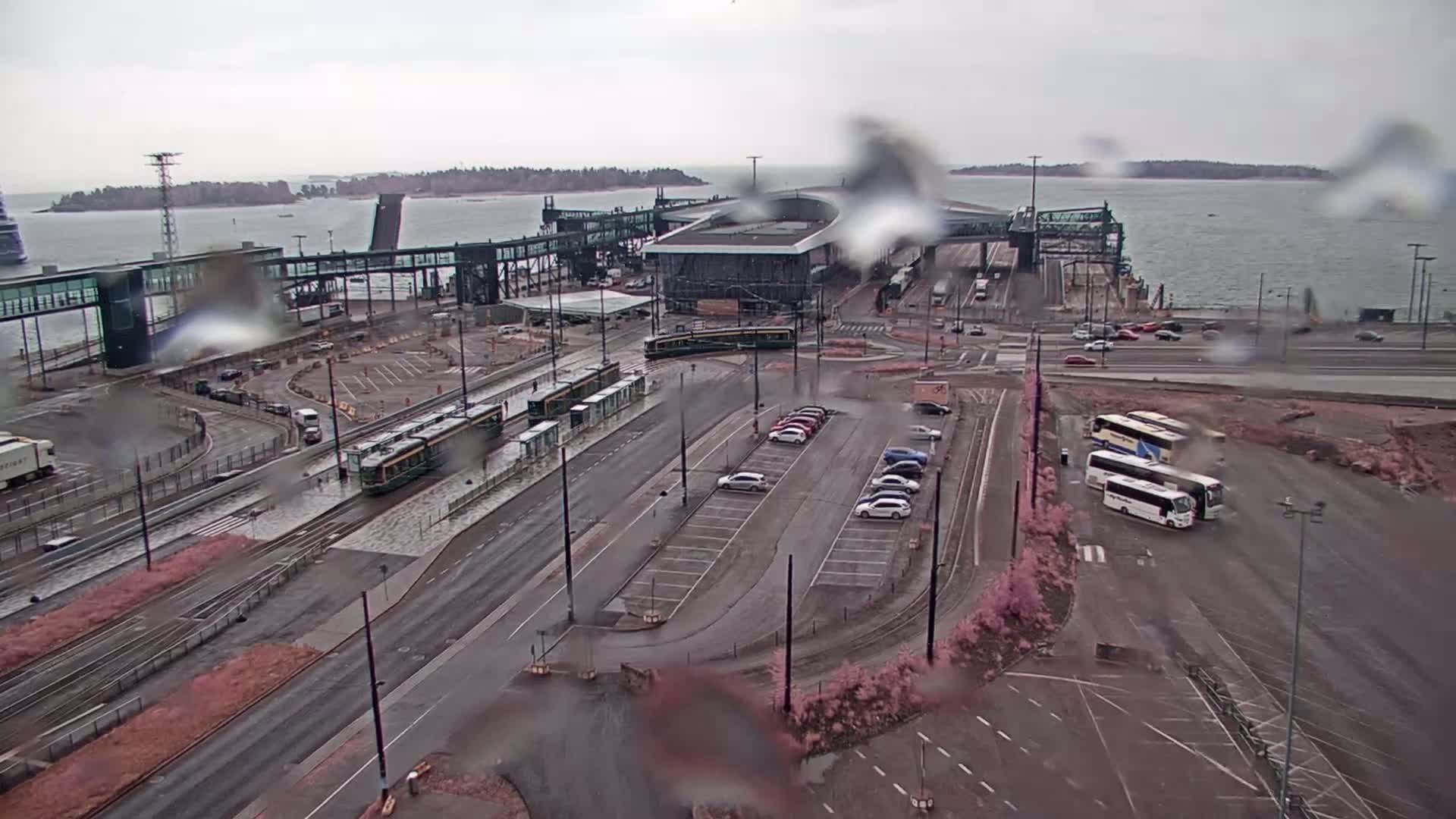 A ferry terminal with trams and buses in a parking lot, under rainy skies, overlooking a body of water.