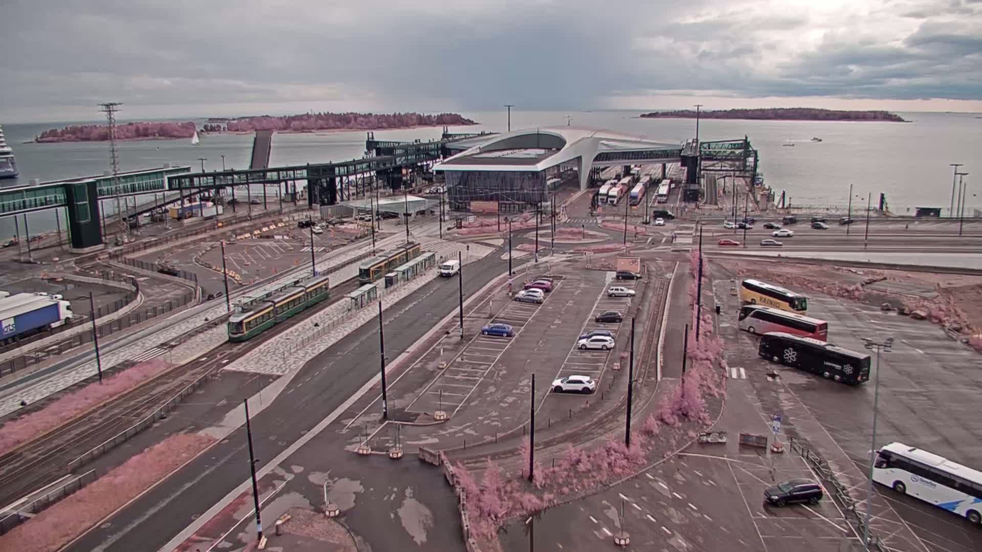 An overcast day reveals a ferry terminal with a tram, cars in parking lots, and buses, all situated near a body of water with islands in the distance.