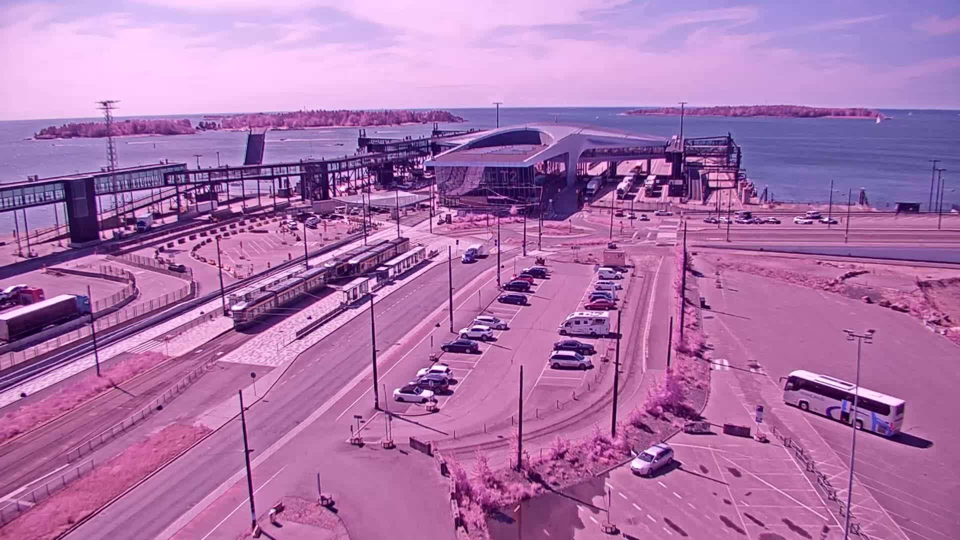 An aerial view shows a ferry terminal with trams and parked vehicles on a sunny day near a body of water with islands in the distance.