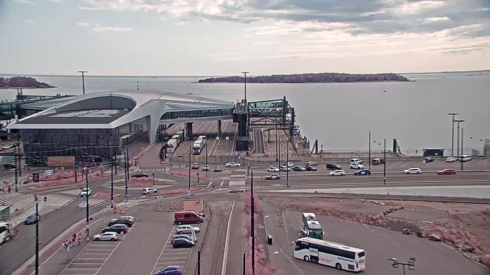 A ferry terminal with a modern building and numerous vehicles, situated on a body of water under a cloudy sky.