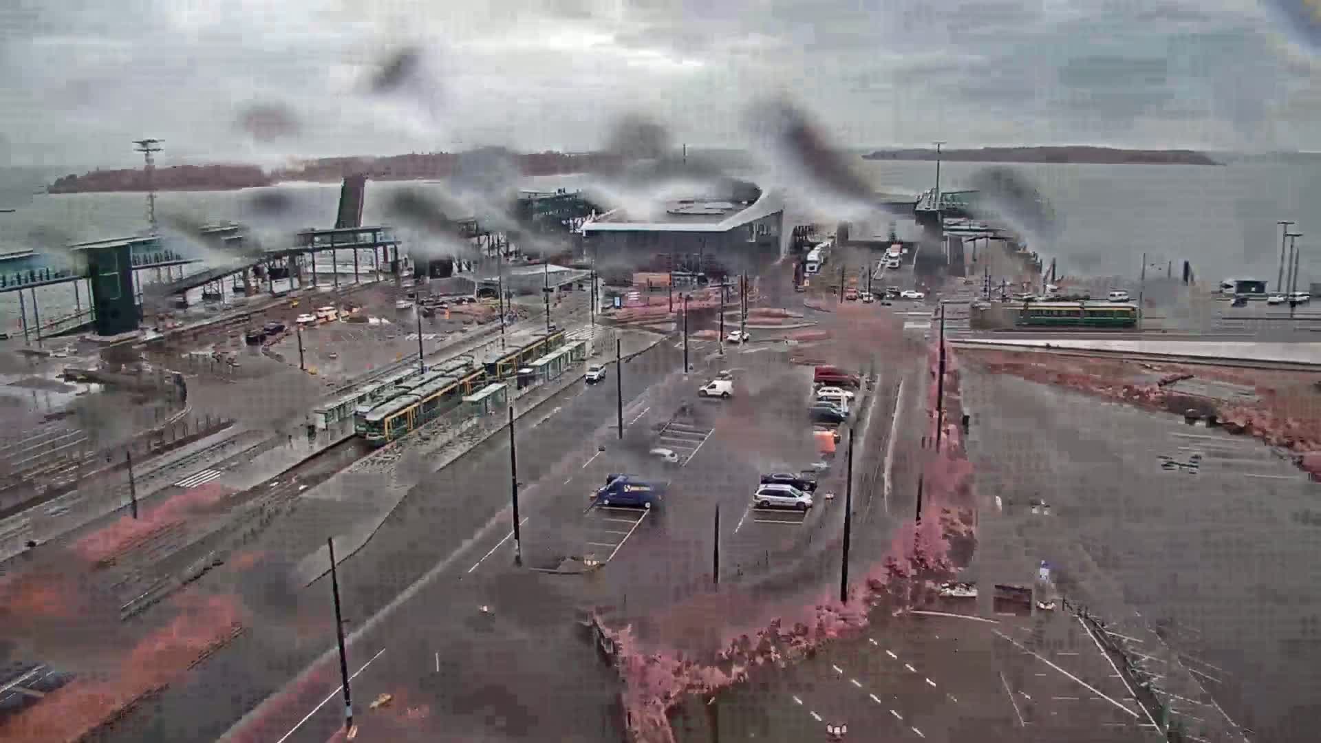A rainy day at a waterfront transit station shows trams and cars parked near a ferry terminal, with a body of water visible in the background.