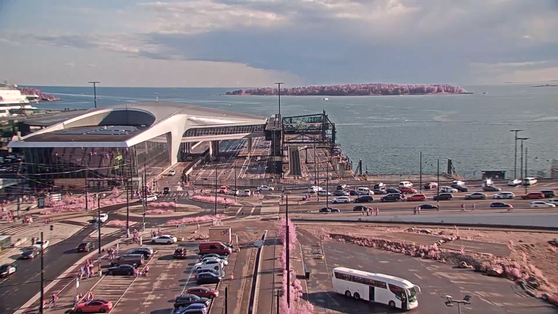 A ferry terminal with a modern building and numerous cars parked around it, overlooks a calm body of water with an island in the distance under a partly cloudy sky.