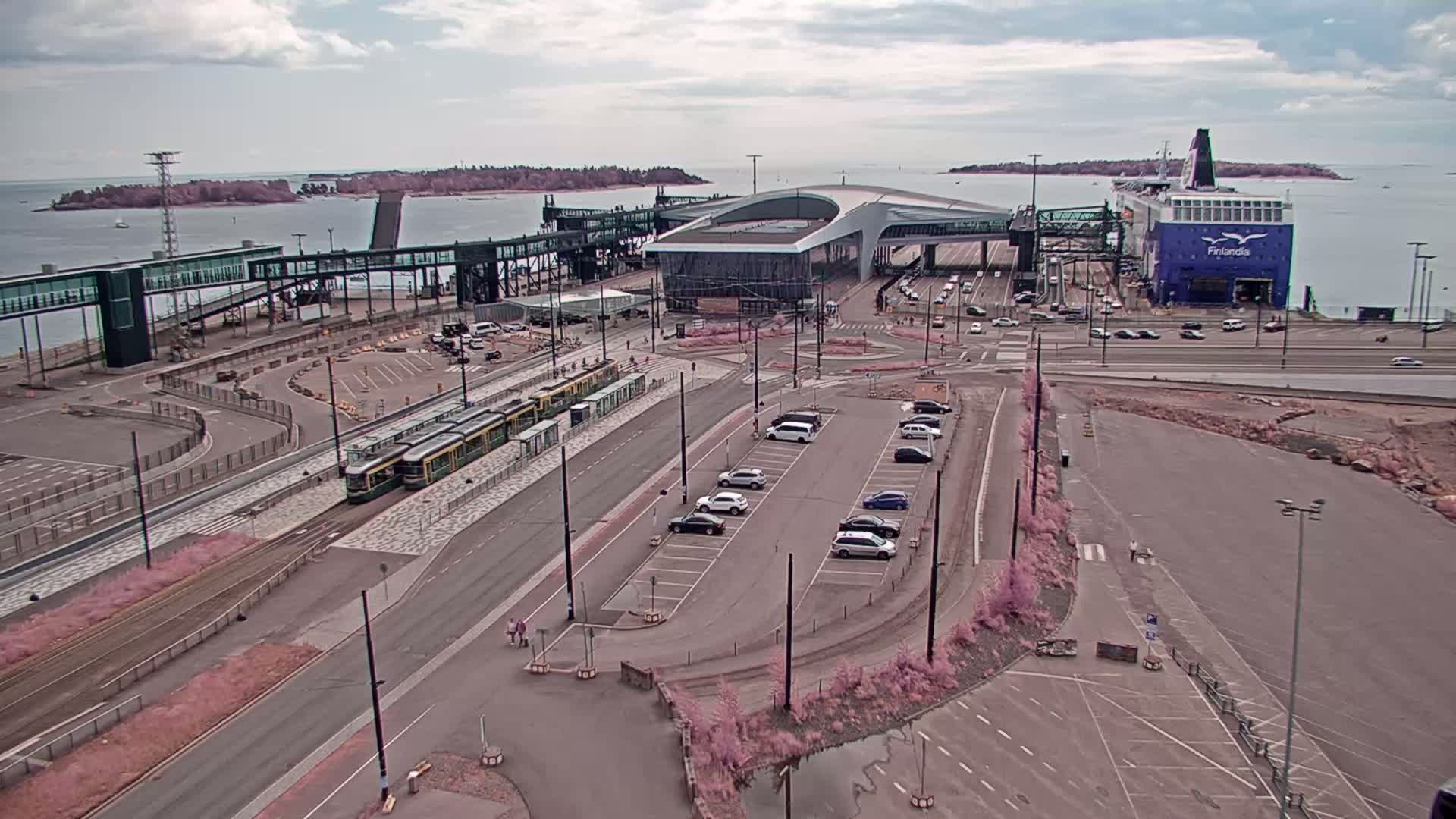 An aerial view shows a ferry terminal with a tram, cars parked in a lot, and a ferry docked under cloudy skies.