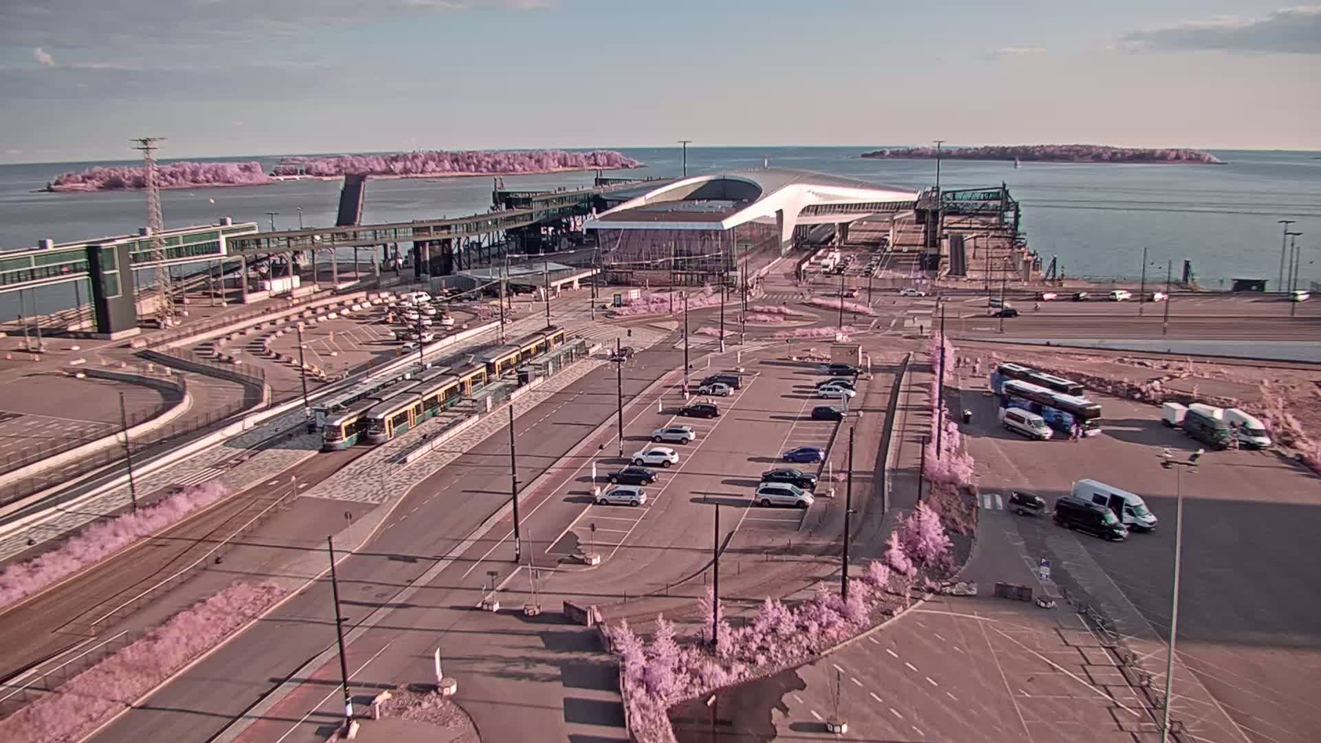 A ferry terminal with a tram, several cars and buses in a parking lot, and a calm body of water with islands in the background under a partly cloudy sky.