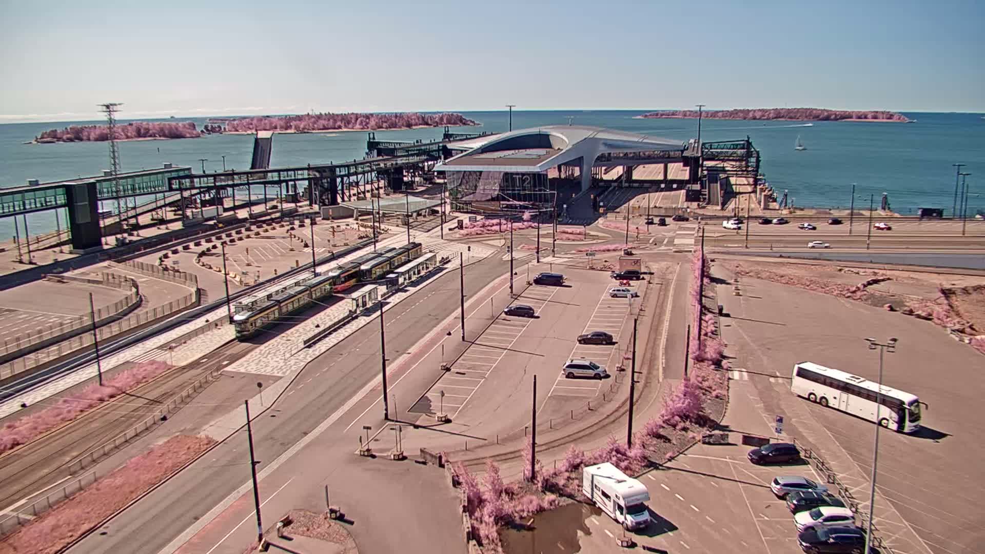 A ferry terminal with several vehicles in parking lots, trams at a station, and a bus, all under a sunny sky with a calm body of water and islands in the background.