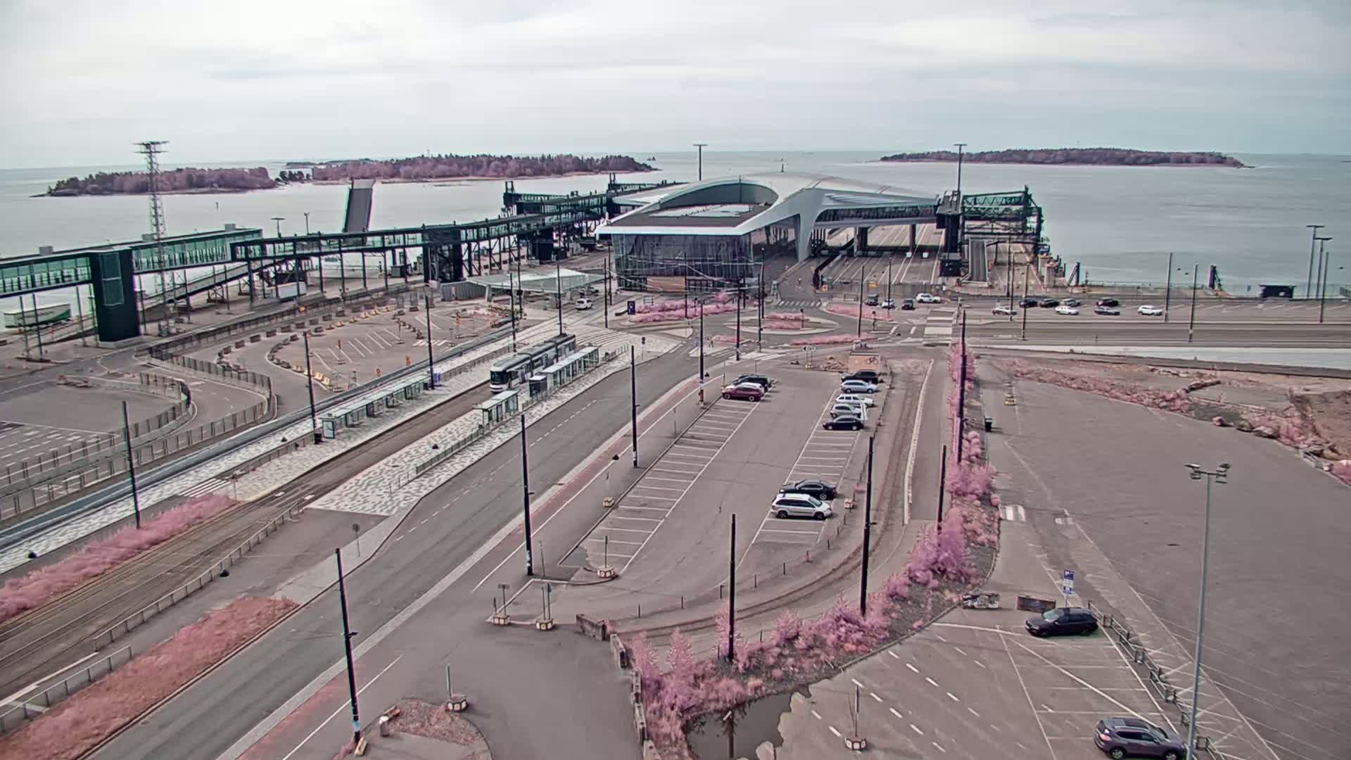 An aerial view shows a ferry terminal with a tram, cars in parking lots, and calm water with islands in the background under an overcast sky.