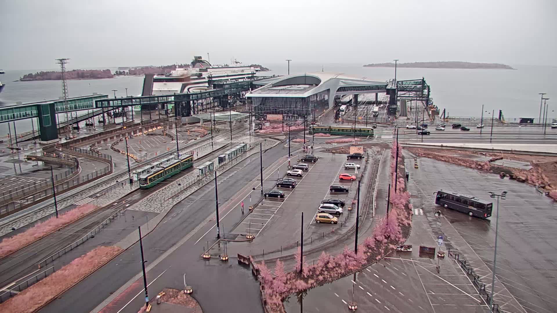A ferry terminal with trams and cars parked in a lot, under an overcast sky on a rainy day.