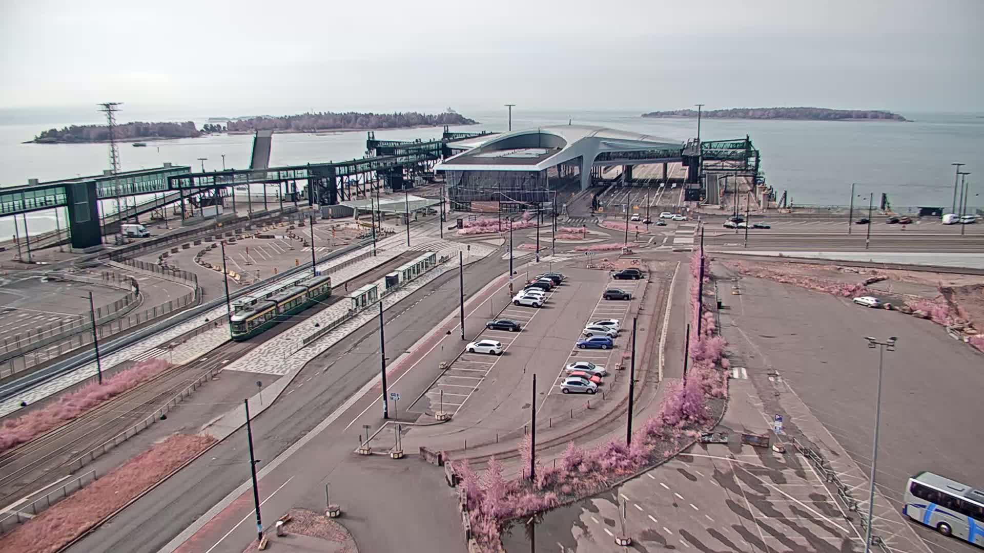 A tram travels along tracks near a ferry terminal and parking lot on an overcast day, with calm water and islands visible in the distance.