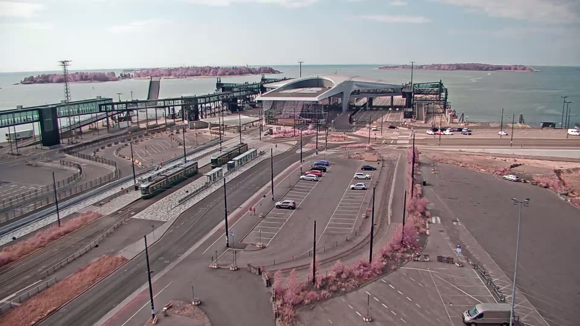 An aerial view shows a ferry terminal with trams, cars in a parking lot, and calm water with islands in the background under a mostly clear sky.