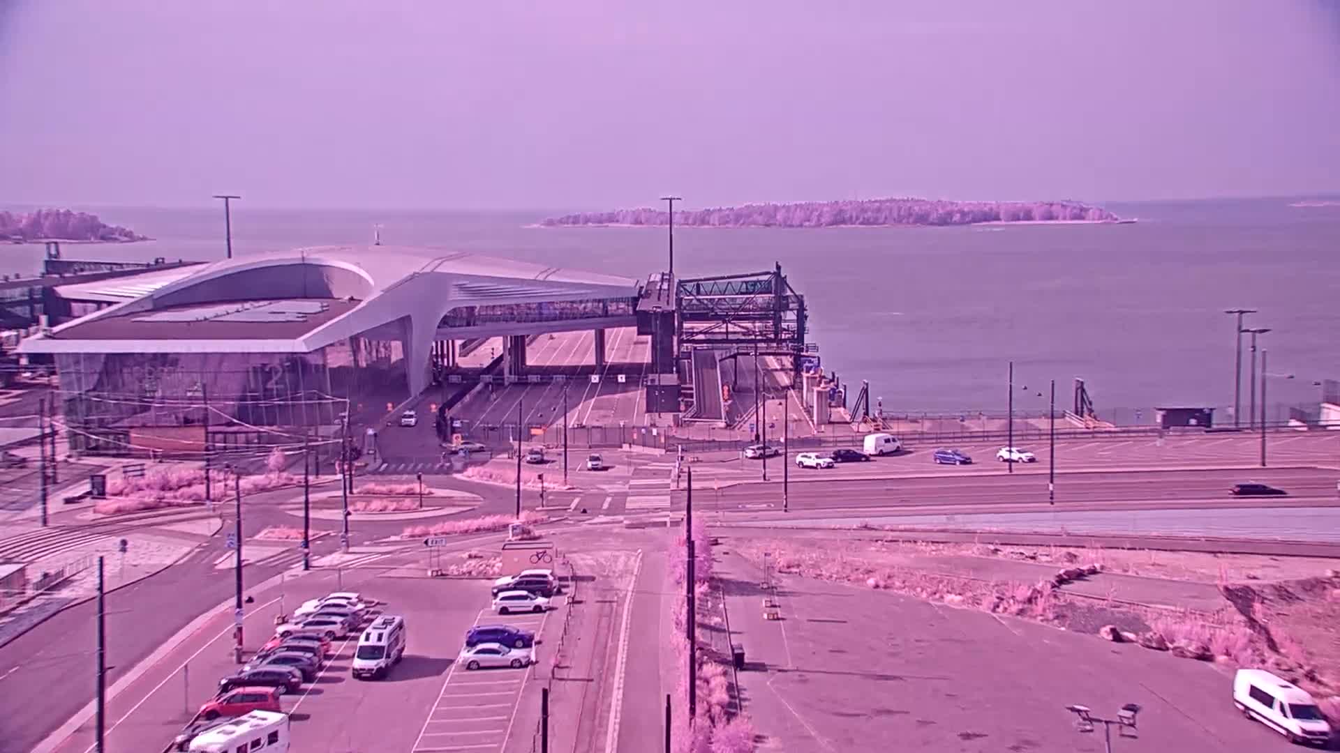 A modern ferry terminal with cars parked nearby overlooks a calm body of water under a hazy sky.