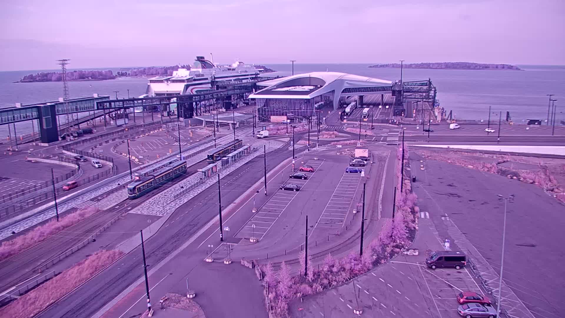 A modern ferry terminal with cars parked nearby overlooks a calm body of water under a hazy sky.