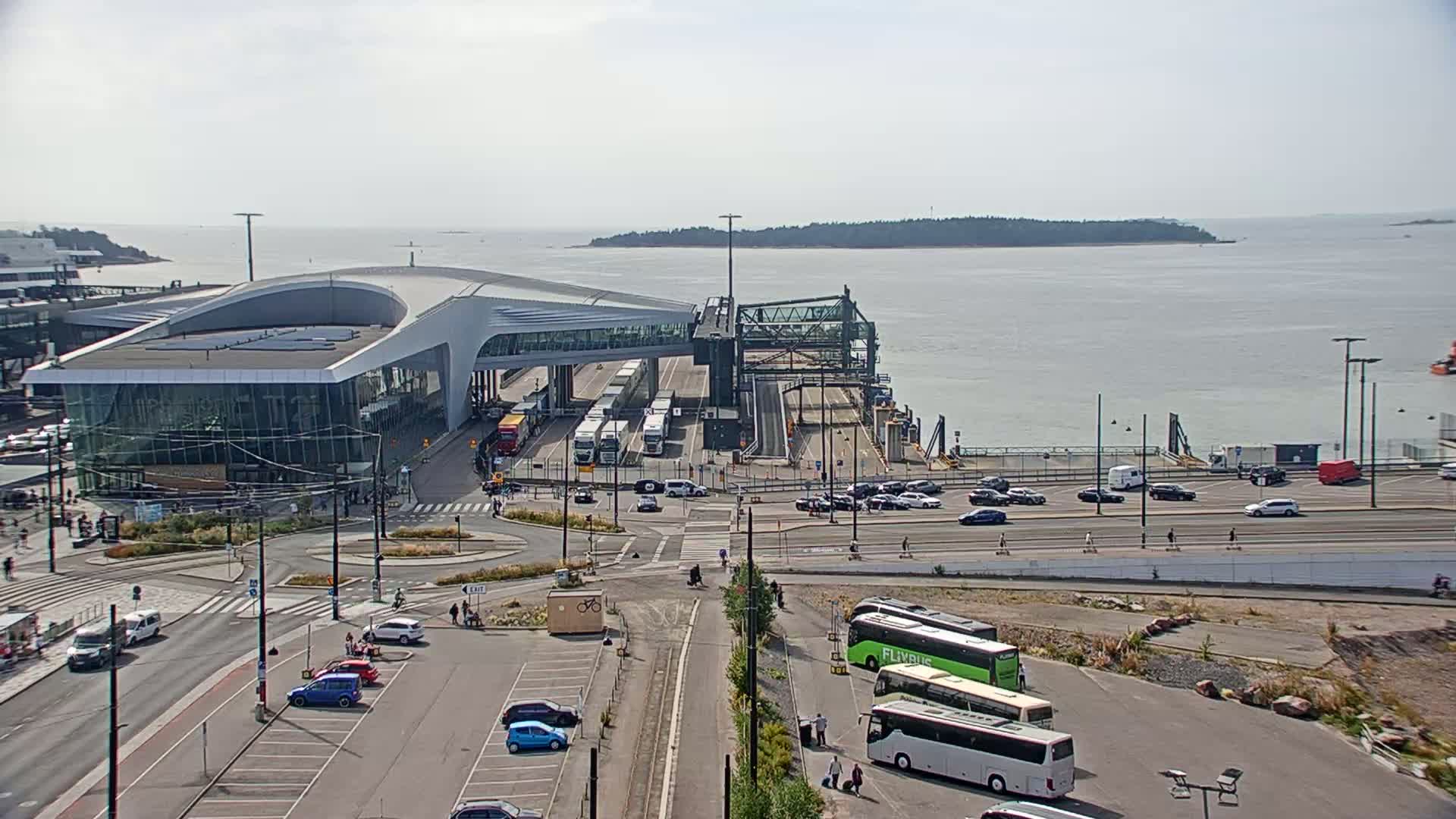 A modern ferry terminal with cars parked nearby overlooks a calm body of water under a hazy sky.