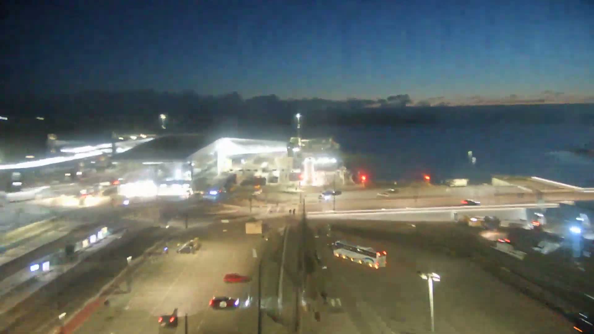 A modern ferry terminal with cars parked nearby overlooks a calm body of water under a hazy sky.