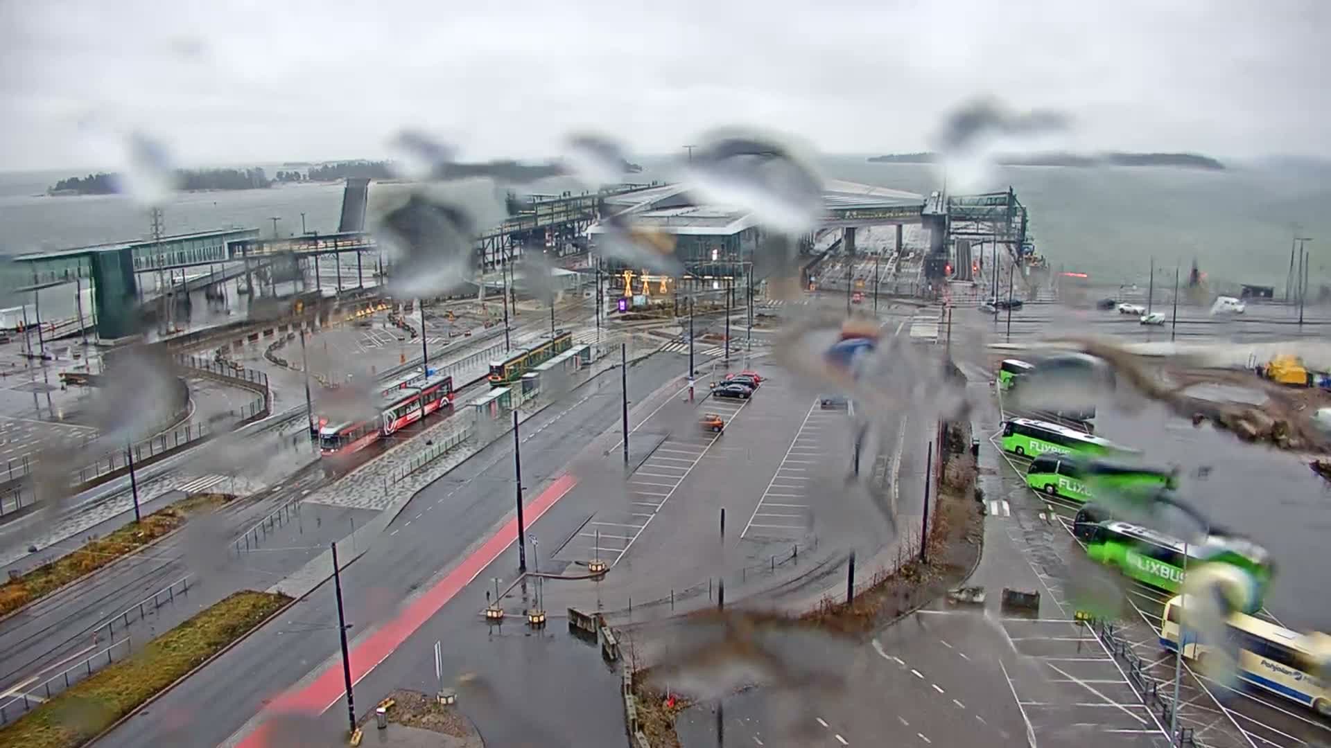 Through a rain-splattered lens, an overcast view reveals a large modern terminal building beside a vast body of water, surrounded by wet roads with a red tram, several green buses, and parked cars.