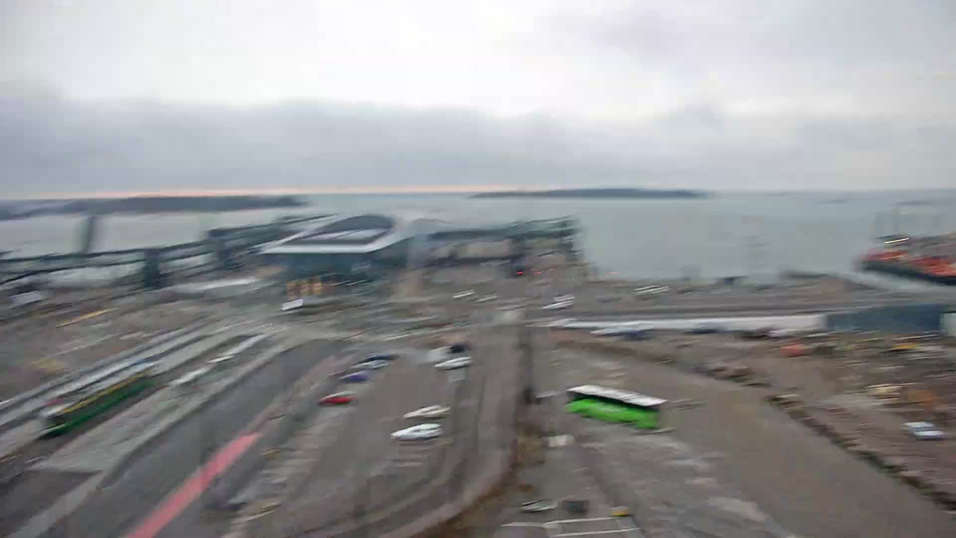 This blurry aerial view depicts an overcast urban port scene featuring a green tram on tracks, multiple vehicles on busy roads, modern waterfront buildings, and distant islands in the gray water.