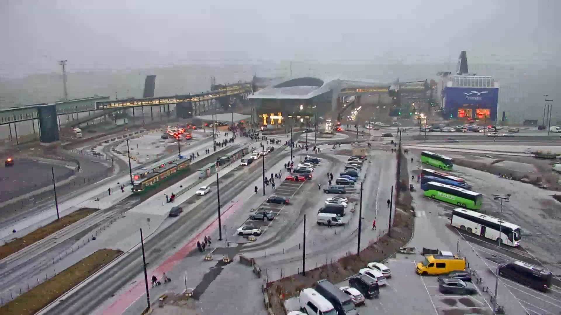This blurry aerial view depicts an overcast urban port scene featuring a green tram on tracks, multiple vehicles on busy roads, modern waterfront buildings, and distant islands in the gray water.