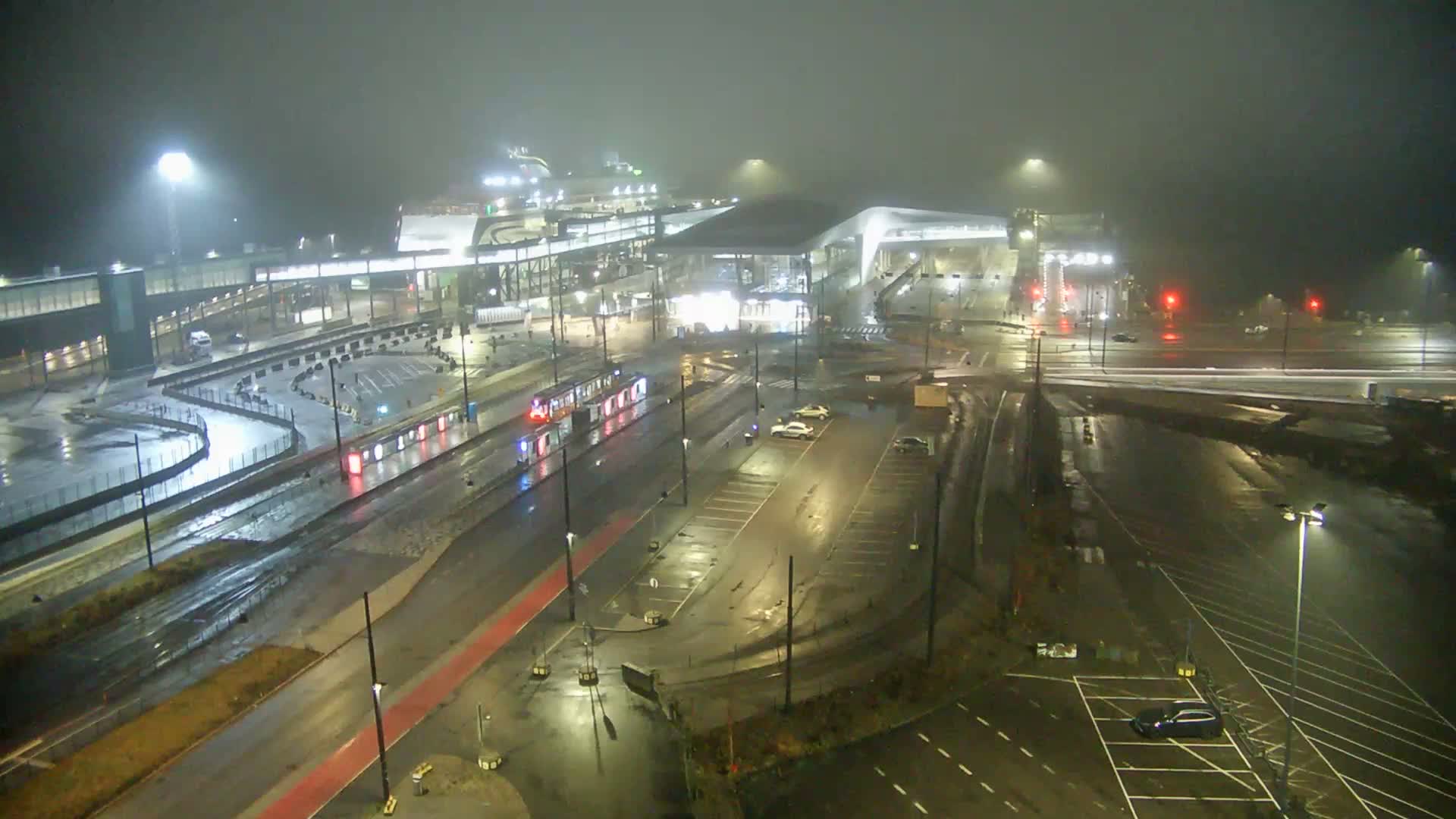 This blurry aerial view depicts an overcast urban port scene featuring a green tram on tracks, multiple vehicles on busy roads, modern waterfront buildings, and distant islands in the gray water.