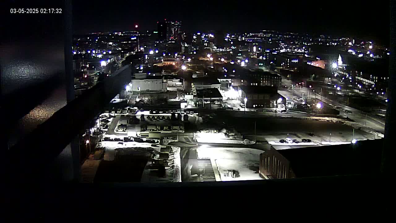 A nighttime aerial view of a city, showing snow-covered ground and illuminated buildings and streets.