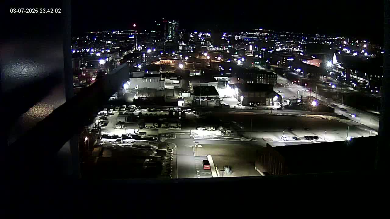 A nighttime, high-angle view of a city's industrial area and surrounding buildings, partially snow-covered, with scattered streetlights illuminating the scene.