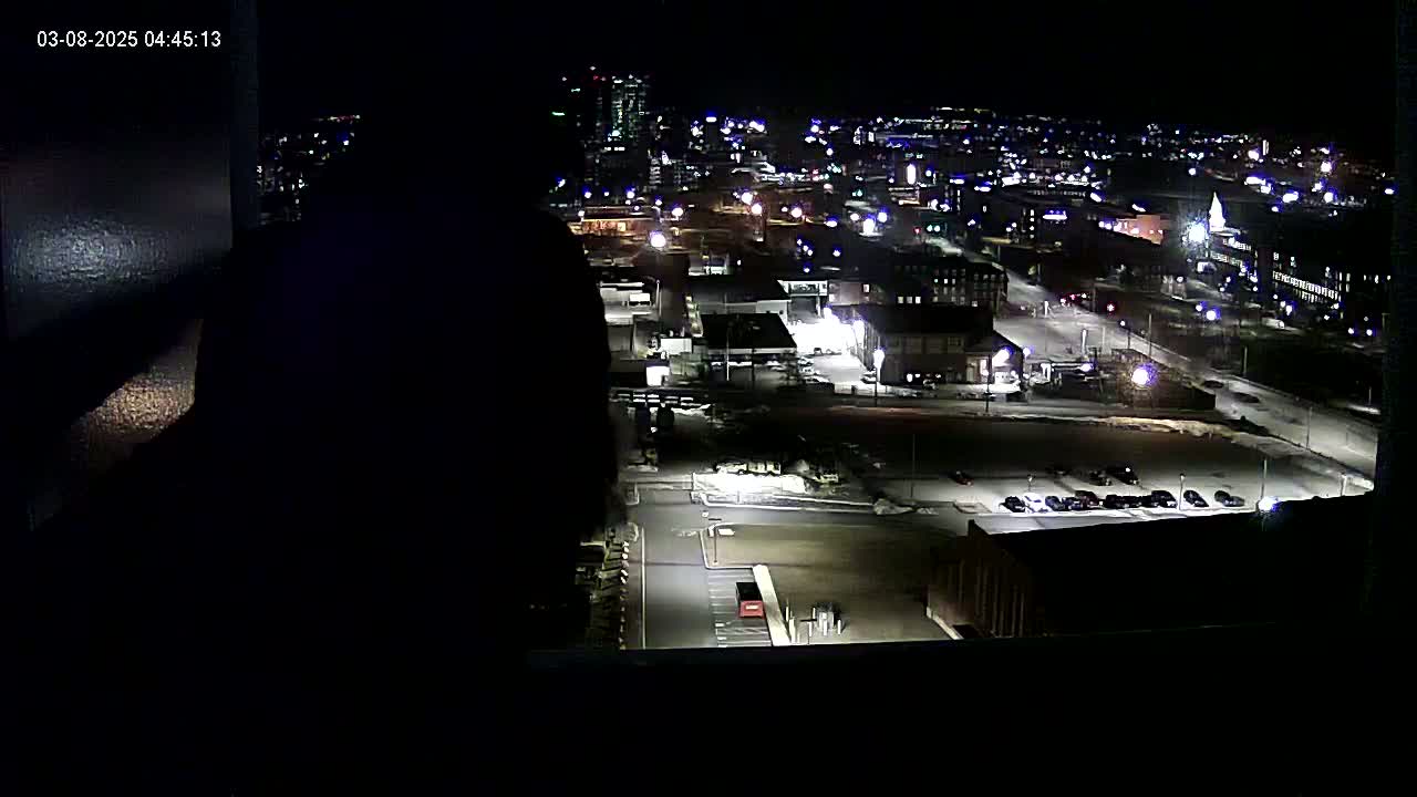 A nighttime view from a window shows a city landscape with a parking lot and several buildings illuminated under clear skies.