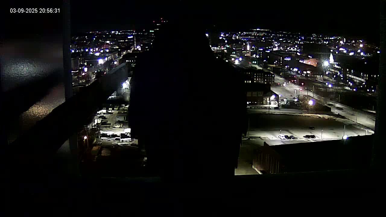 A nighttime, high-angle view of a city illuminated by streetlights and building lights, with patches of snow or ice visible on the ground.