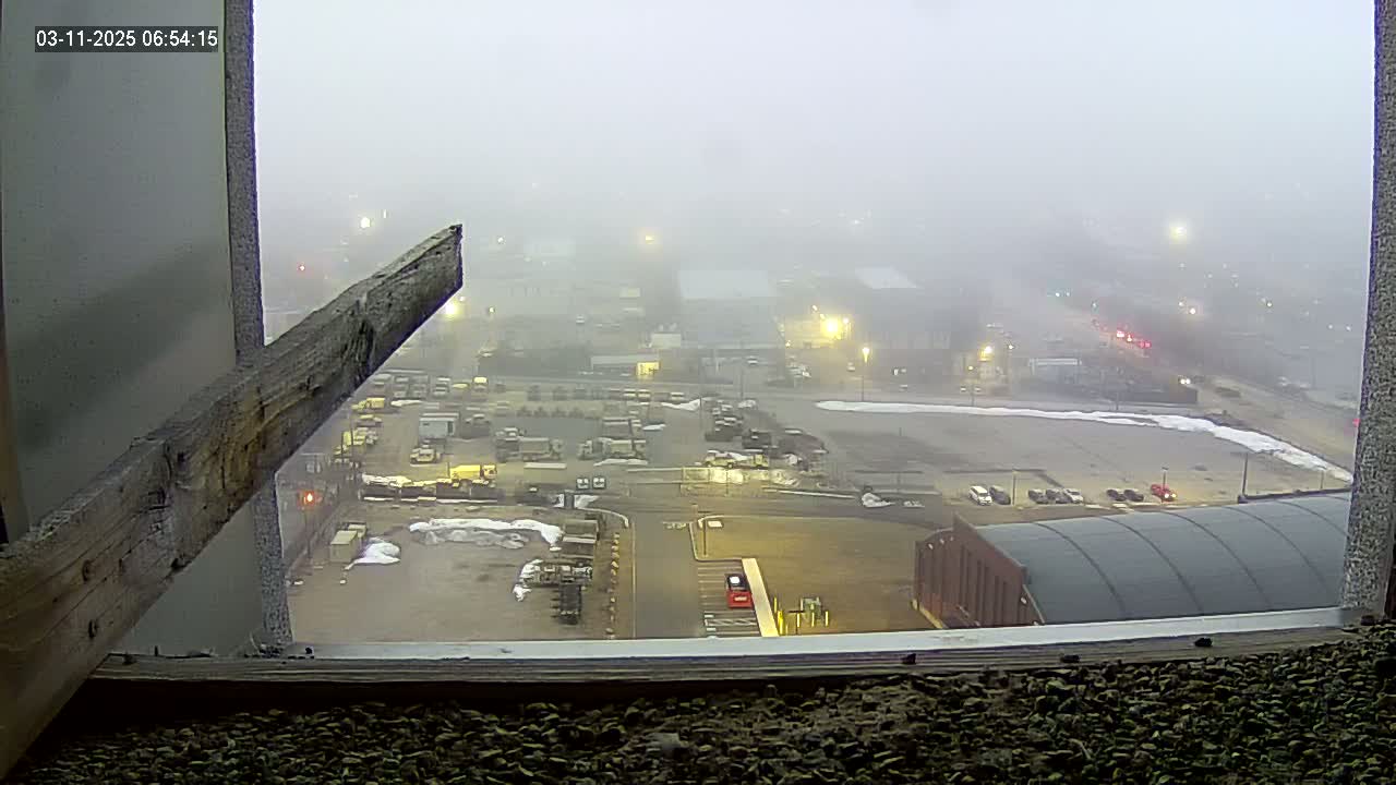 A foggy view from a window shows a large industrial area with parked vehicles, buildings, and patches of snow on the ground.