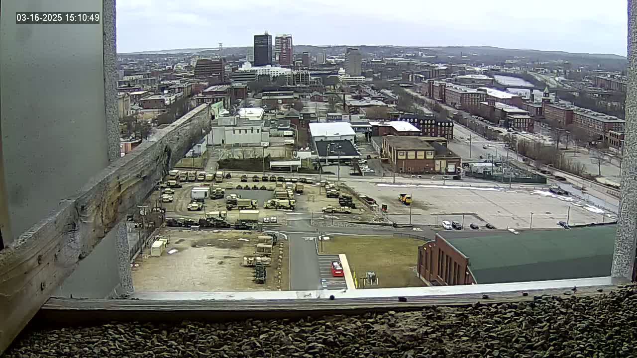 A high-angle, long shot on an overcast day shows a large parking lot filled with military vehicles next to an industrial area and a city in the distance.