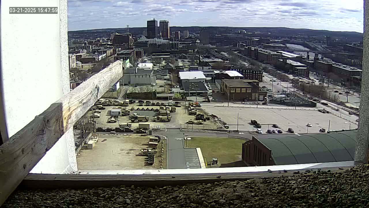 A high-angle view on a partly cloudy day shows a city with a large parking lot full of military vehicles, surrounded by buildings and a river in the distance.
