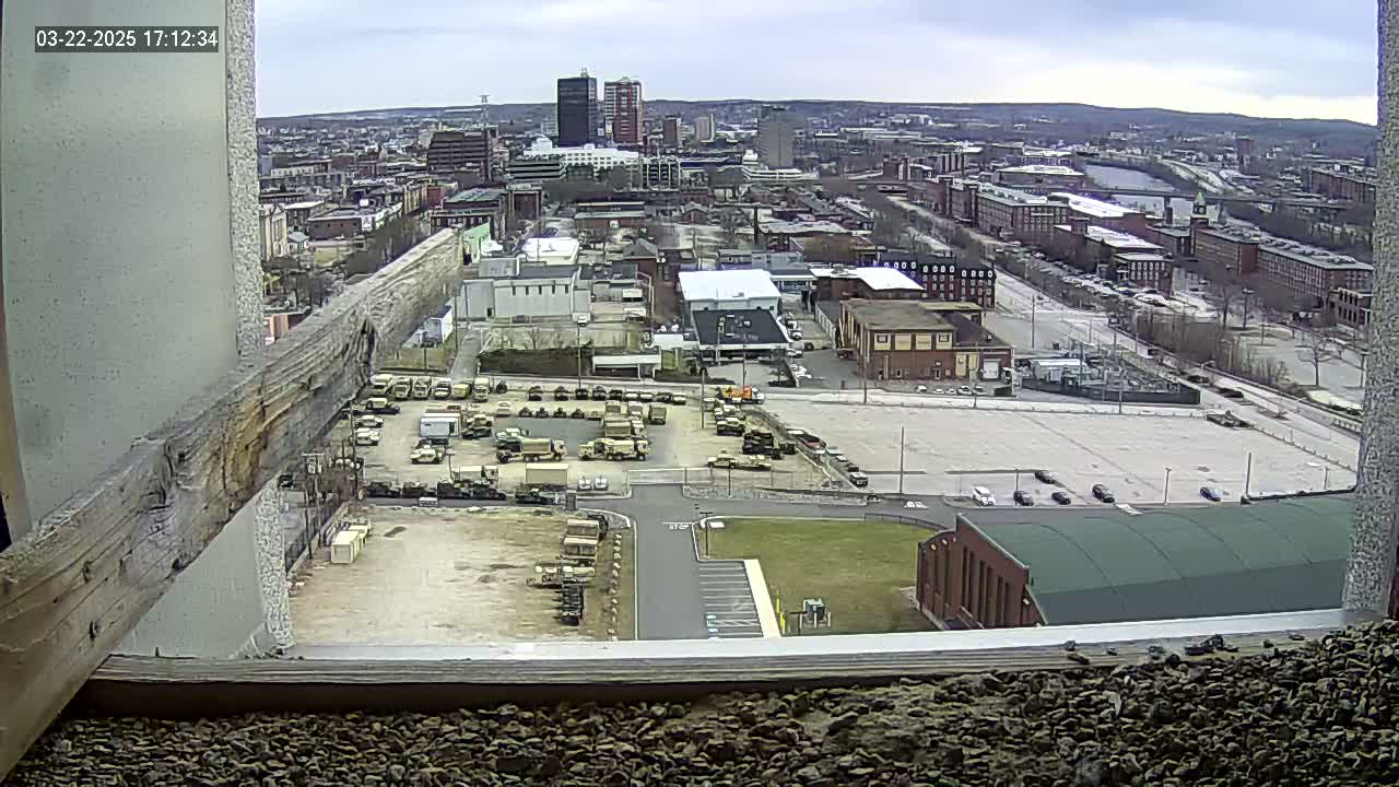A high-angle, outdoor view on an overcast day shows a large parking lot filled with military vehicles next to a city with numerous buildings and a river in the distance.