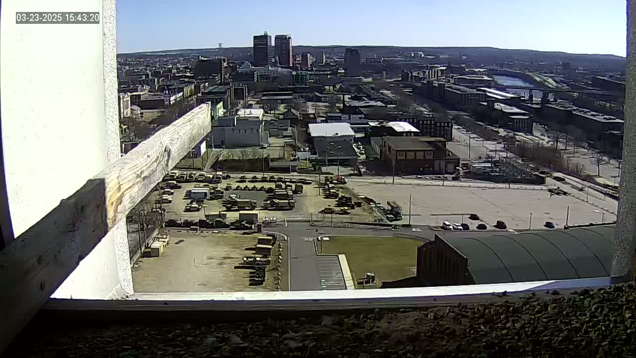 A high-angle, long shot on a sunny day shows a cityscape with a large parking lot full of military vehicles, and a river in the distance.