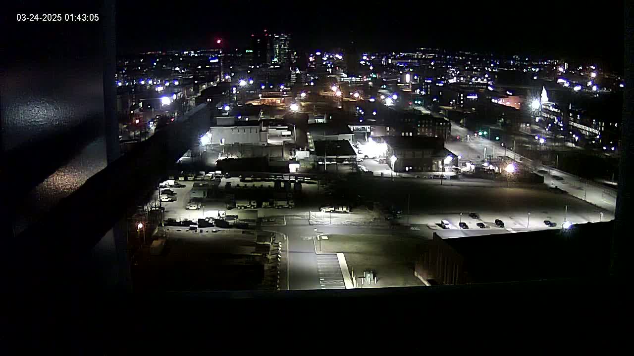 A nighttime aerial view of a city's industrial area, showing illuminated buildings, parking lots, and roads, under a clear, dark sky.