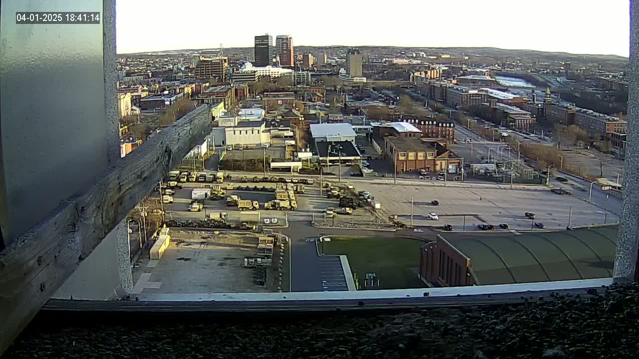 A high-angle, long shot of a city under a clear, twilight sky shows a large parking lot filled with military vehicles near industrial buildings and a river.