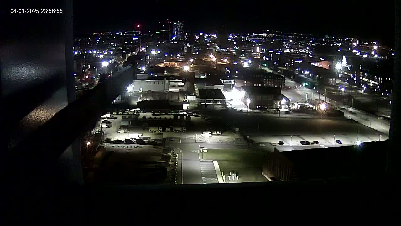 A nighttime, high-angle view of a city illuminated by streetlights and building lights, showing parking lots, roads, and buildings.