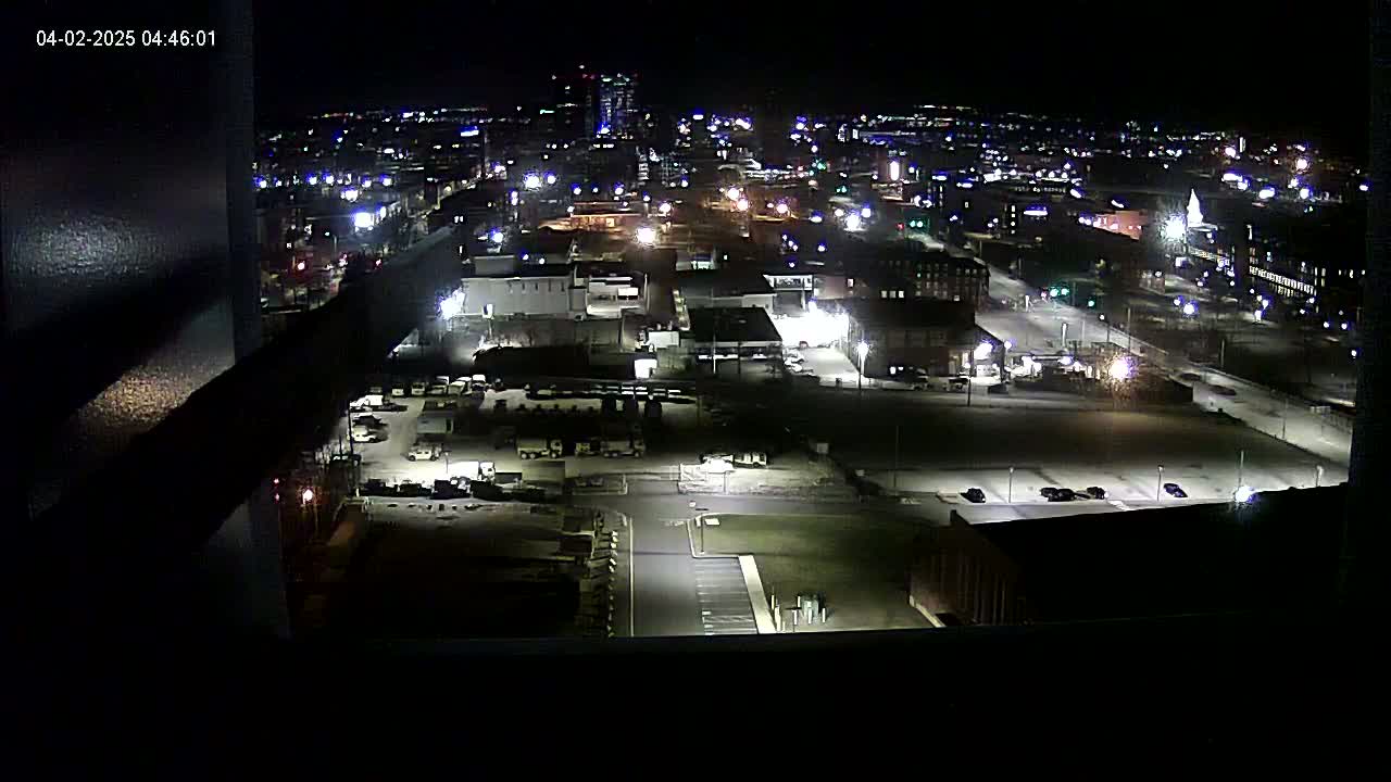 A nighttime, high-angle view of a city's industrial area with sparsely parked cars, illuminated by streetlights and building lights.